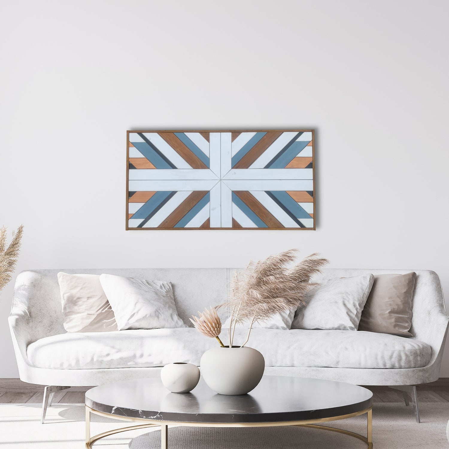 Handcrafted geometric wood wall art featuring a starburst pattern in soft blue, white, and natural wood tones, mounted above a modern white velvet sofa in a bright, minimalist living room with pampas grass decor and neutral accents.