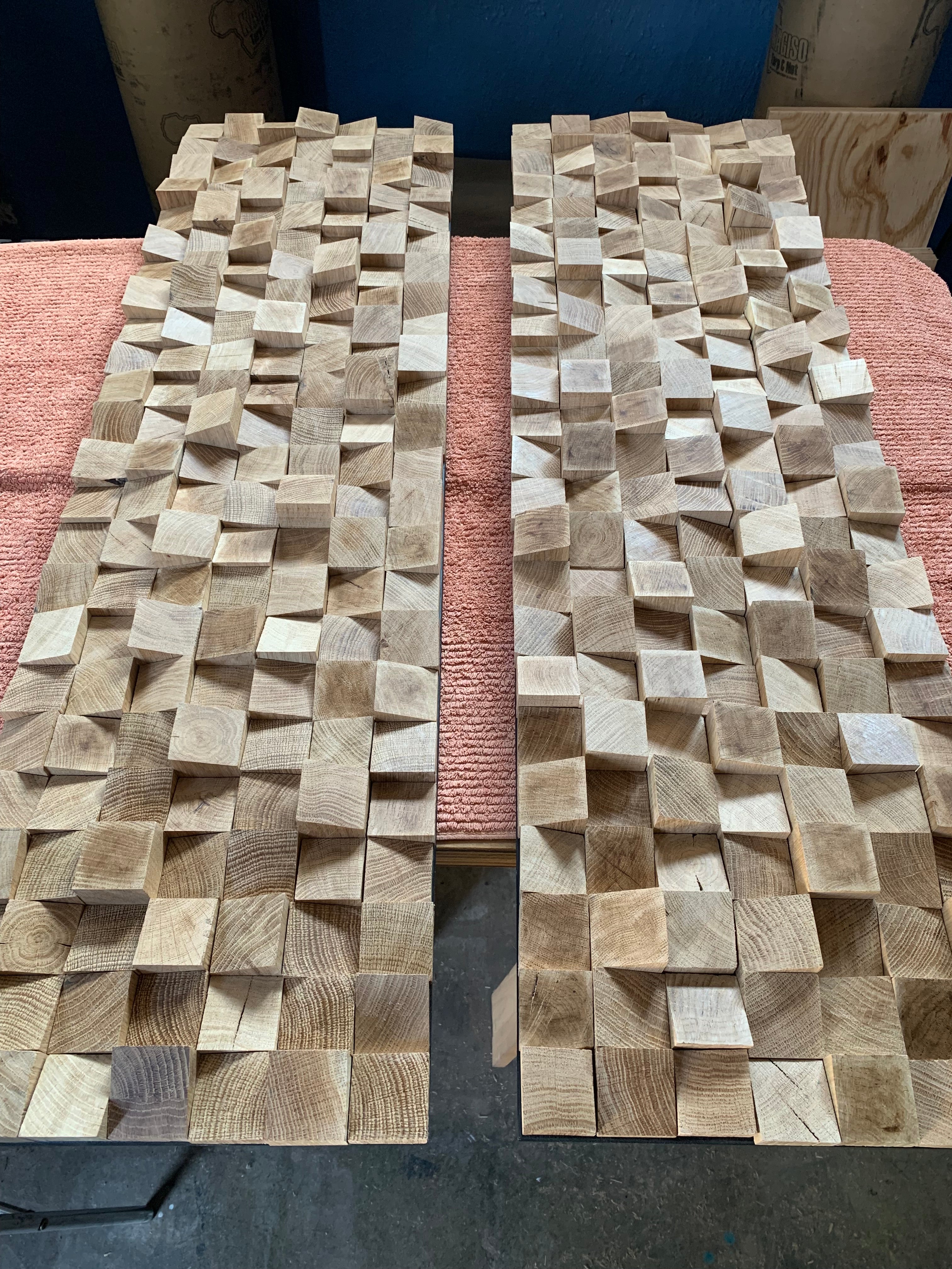 Two handmade sculptural wall panels crafted from white oak blocks with angled surfaces and organic depth, shown resting on a workshop bench.
