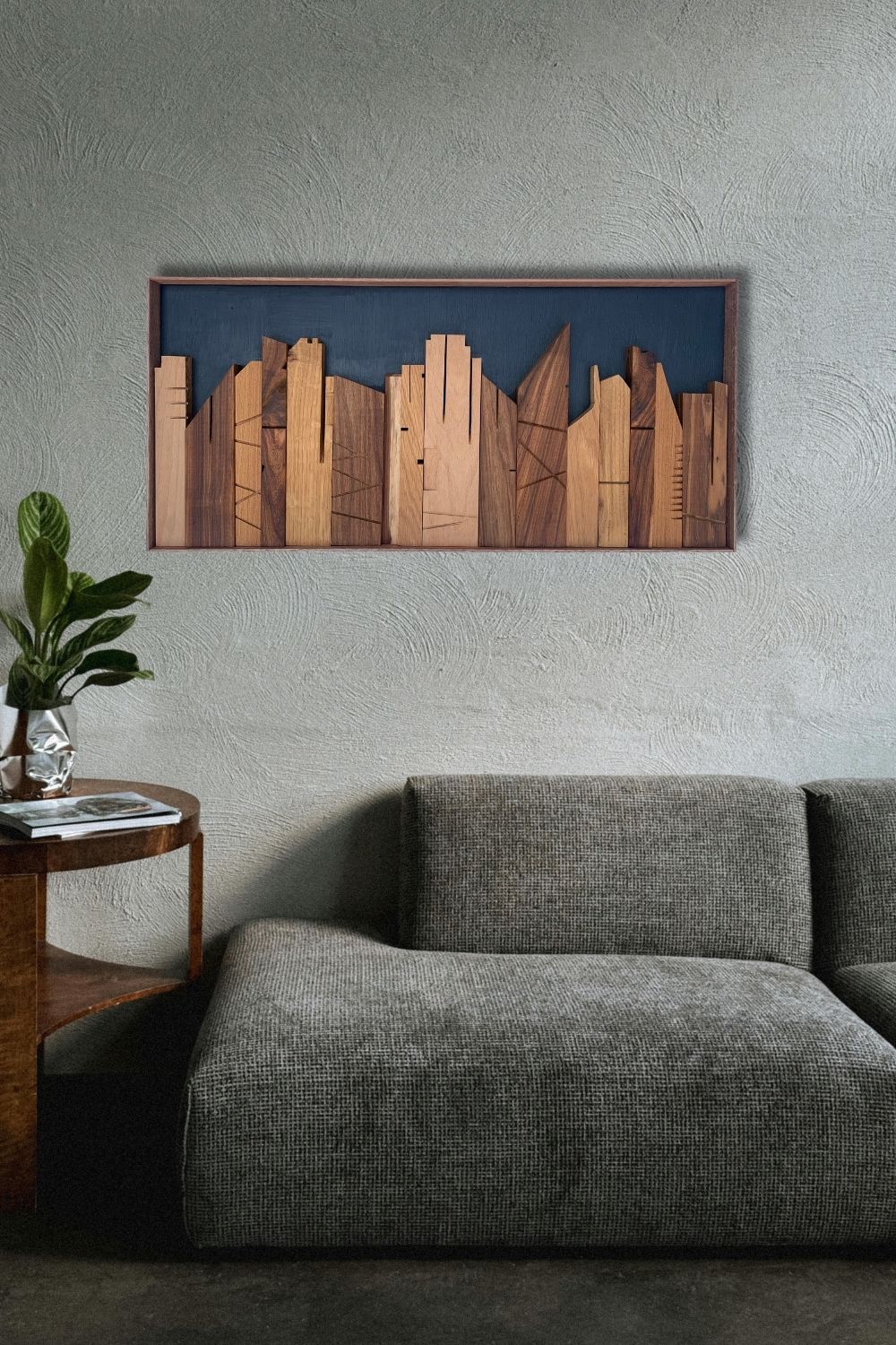 Cityscape wooden wall art displayed above a grey sofa on a textured wall, with layered timber skyline forms and a deep blue backdrop.
